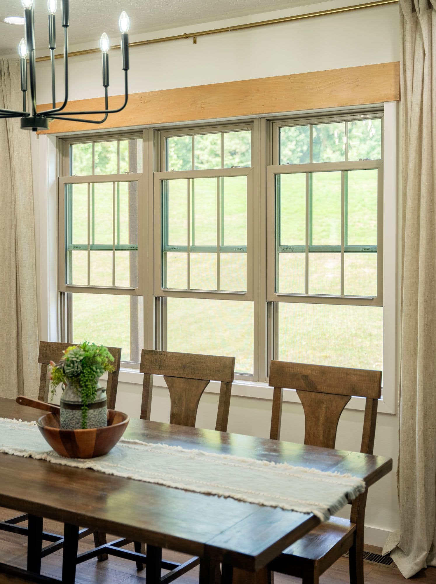 A cozy dining room with a wooden table, rustic chairs, and a white cloth runner. Bright, large windows reveal a lush green lawn outside, under a modern black chandelier.