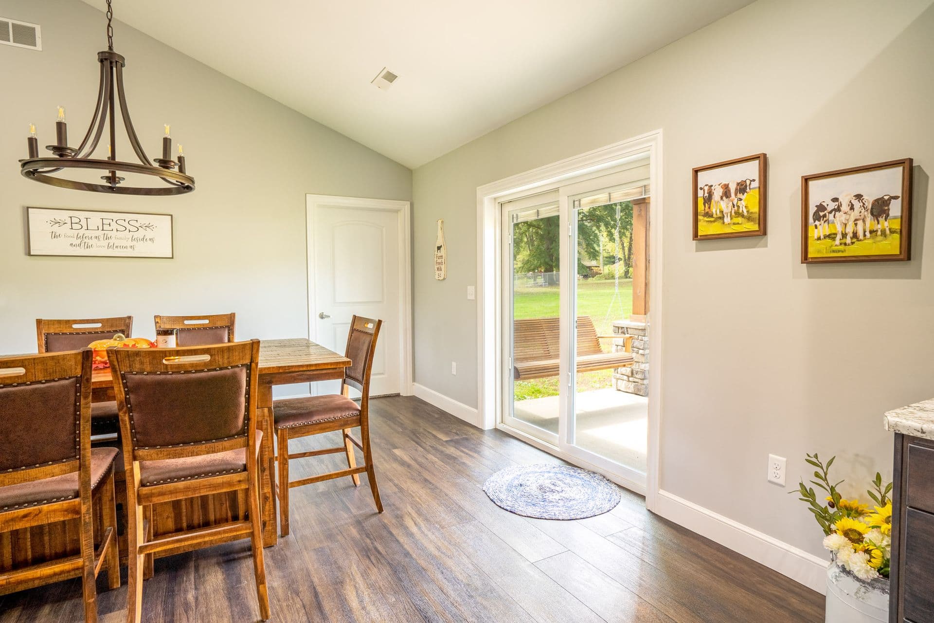 Dining room with a wooden table and chairs, lit by a chandelier. A "Bless" sign hangs on a gray wall. Sliding doors open to a green yard. Cozy and welcoming.