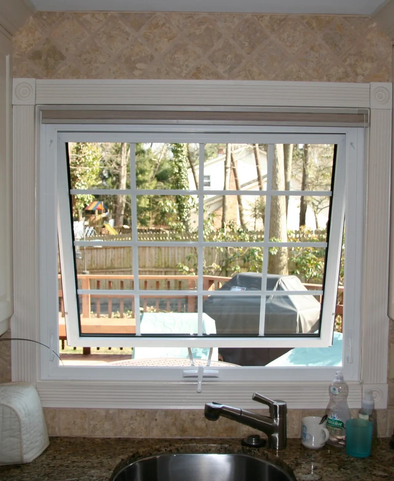 Kitchen window ajar above a sink, showing a deck with a grill and greenery outside. The scene feels calm and domestic, with soft natural light.