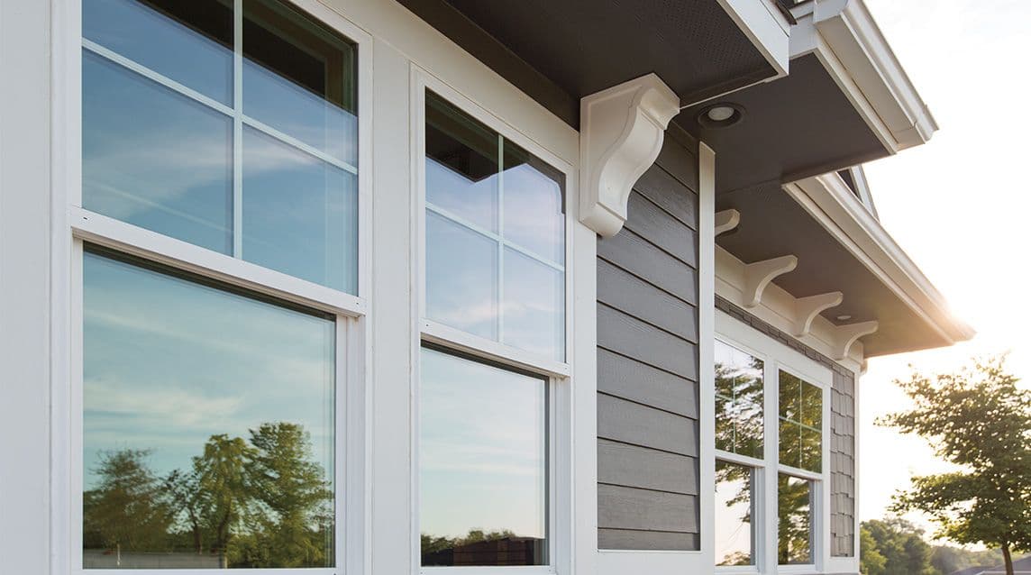Close-up of a modern house exterior, highlighting white-framed windows and gray siding. Sunlight casts warm tones, reflecting trees in the glass.