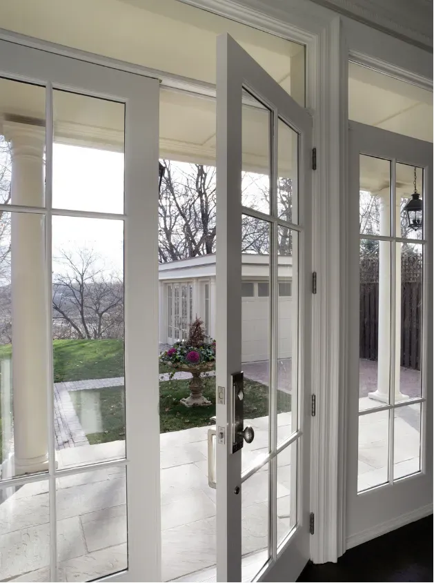 Open French doors reveal a sunlit patio with a lush garden, white columns, and a distant view of a garage. The scene exudes a peaceful, inviting ambiance.