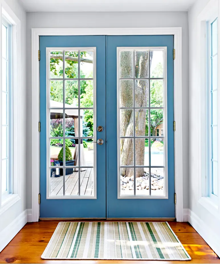 French doors with blue frames and glass panels open to a sunny backyard, revealing a large tree and garden. A striped rug lies on wooden floors inside.