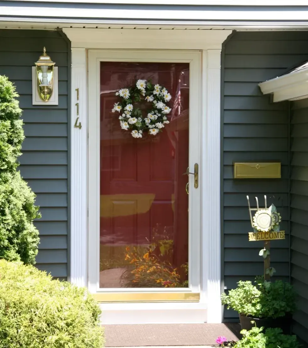 A red front door with a glass storm door features a daisy wreath. The door has house numbers "114" and is surrounded by blue siding. Nearby, a "Welcome" sign is visible, flanked by greenery, creating a welcoming, homey vibe.