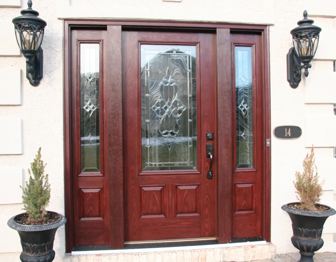 Elegant wooden front door with intricate glass panels, framed by two lantern-style lights. Two potted plants add a welcoming feel.