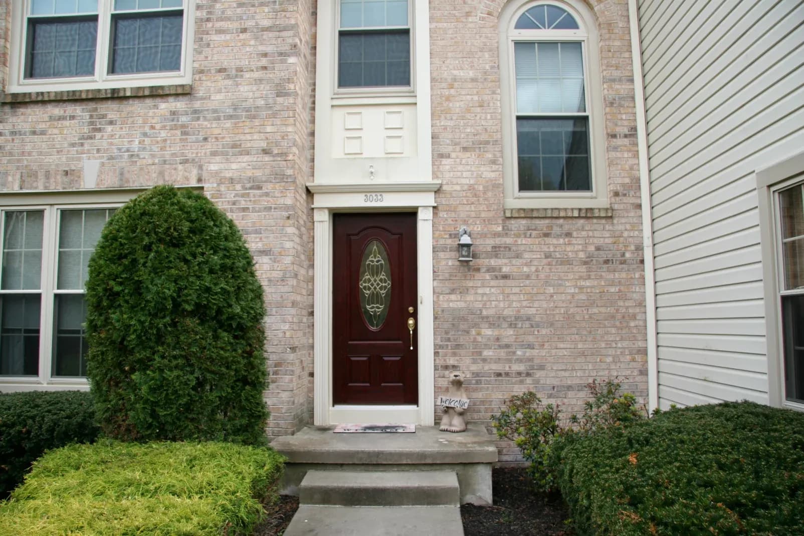 Brick house entrance with a dark wooden door featuring ornate glass. Symmetrical windows frame the door, surrounded by green shrubs, conveying a welcoming tone.