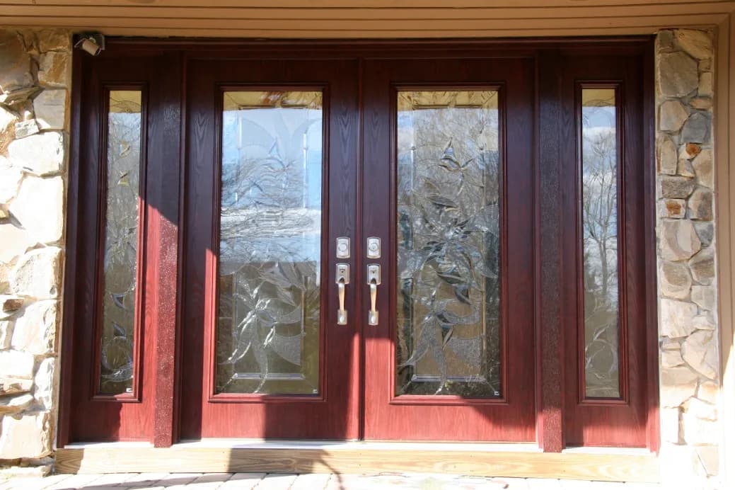 Elegant brown double doors with decorative frosted glass panels, framed by stone walls. Sunlight casts shadows, creating a warm, welcoming tone.