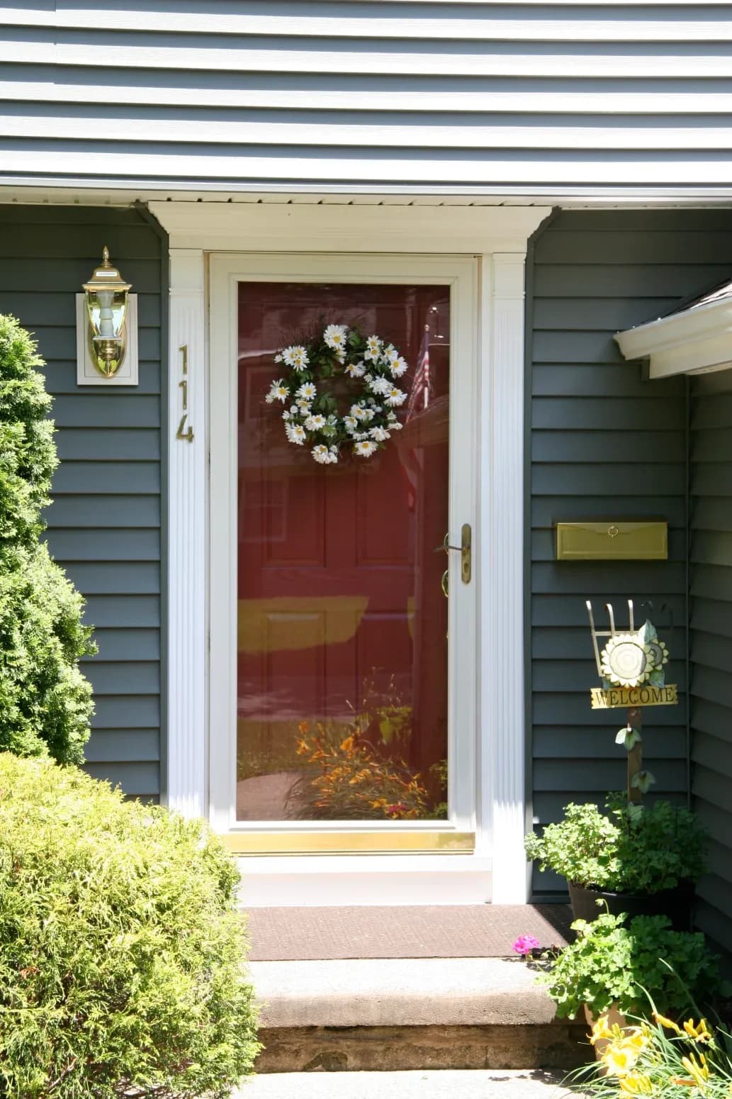 Front door of a house with a glass storm door and a red inner door, adorned with a daisy wreath. A welcome sign and potted plants convey warmth.