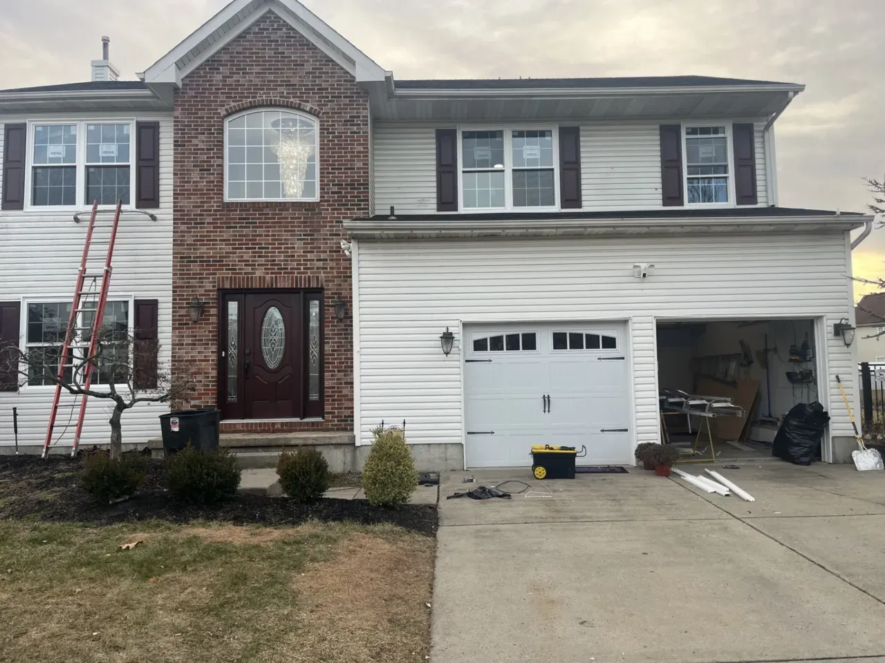 Two-story suburban house with a red brick front, large windows, and a white garage door. A red ladder leans against the house, suggesting ongoing repairs.