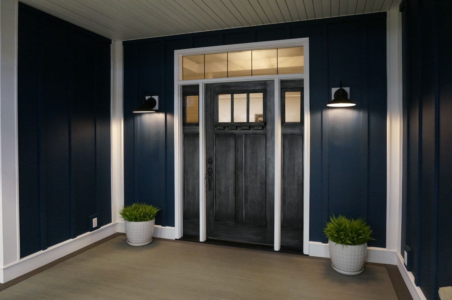 A modern entrance with a dark wood door, flanked by glass panels, set against navy siding. Two potted plants and wall sconces add symmetry and warmth.