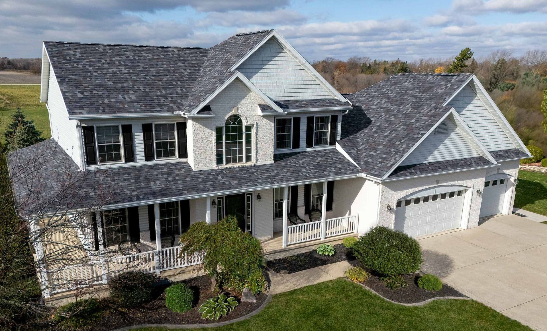 Two-story white farmhouse with gray shingle roof, front porch with white railings, two-car garage, and landscaped front yard with shrubs and lawn