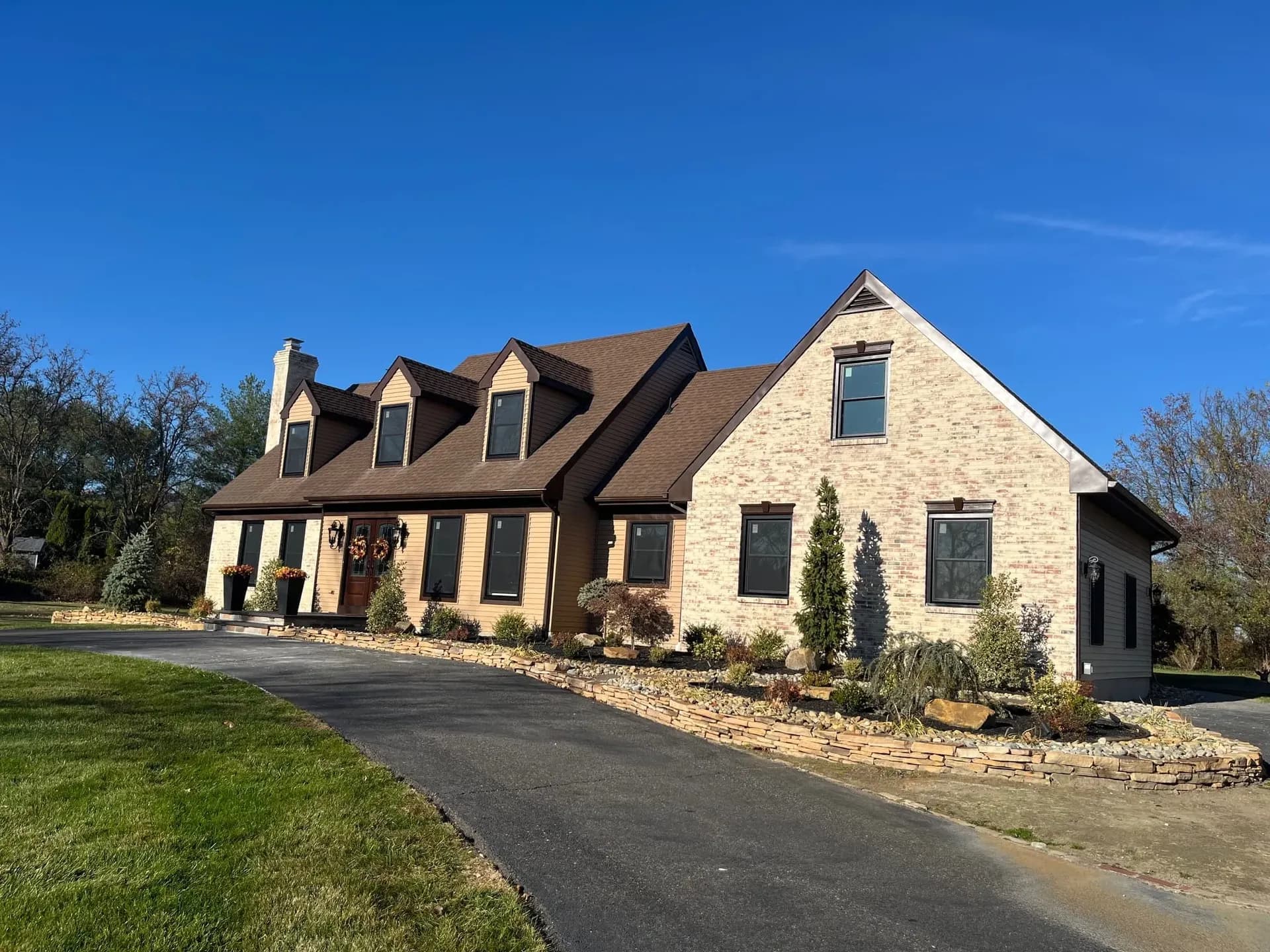 A charming two-story house with a sloped brown roof and beige brick facade. It features dormer windows and a tidy garden. The sky is clear and blue.