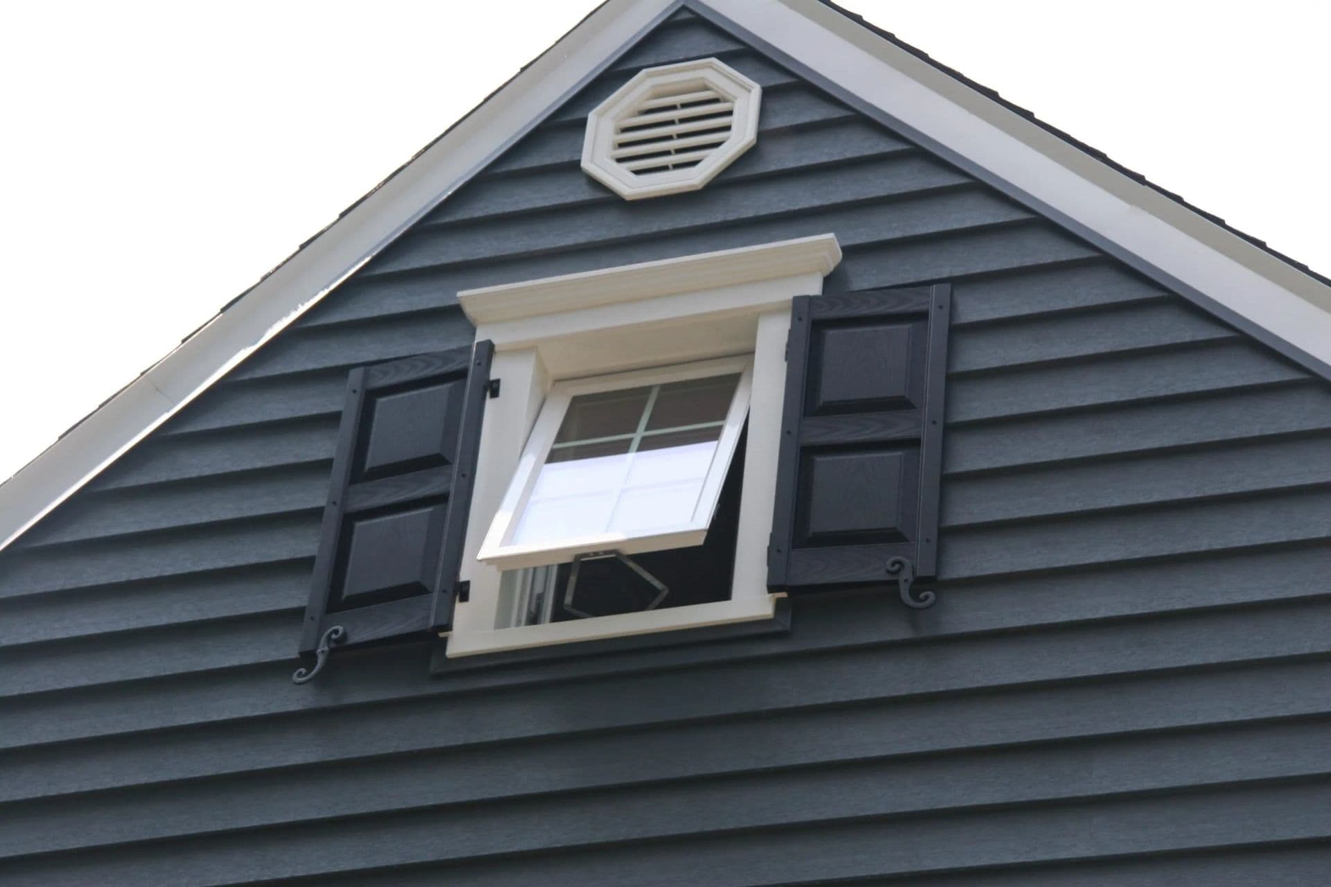Gabled roof with a small, open window featuring black shutters and white trim. The blue siding adds contrast, creating a crisp, classic look.