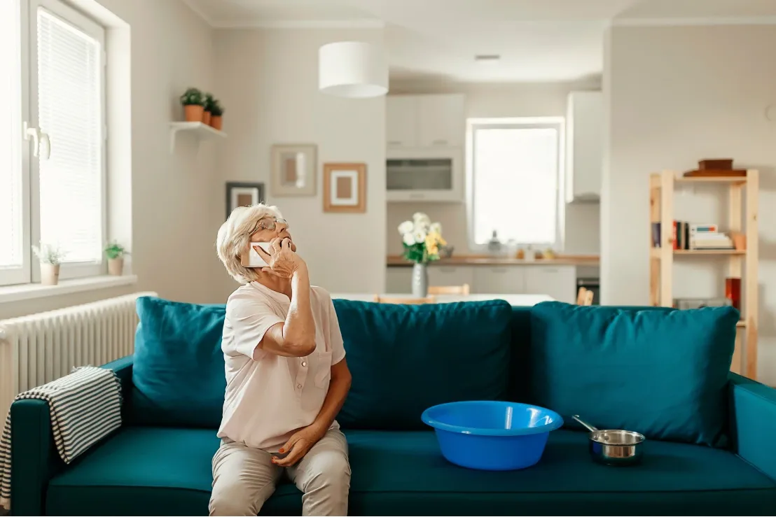 Elderly woman sitting on a teal sofa, speaking on the phone, looking concerned. A blue basin and pot sit beside her, suggesting a possible leak.