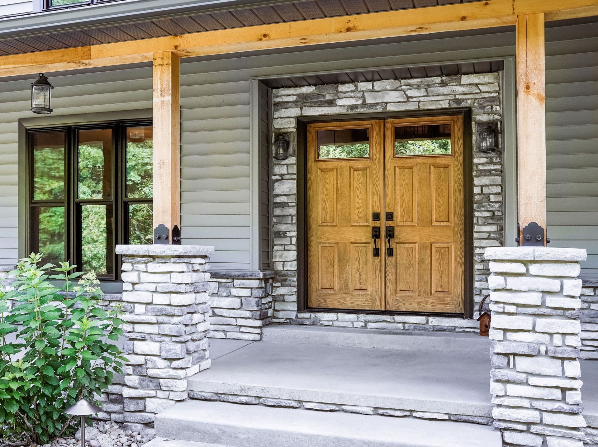 Front porch with double wooden doors framed by rustic stone, flanked by large windows. Wooden columns support a covered gray siding exterior. Lush greenery adds vibrancy.