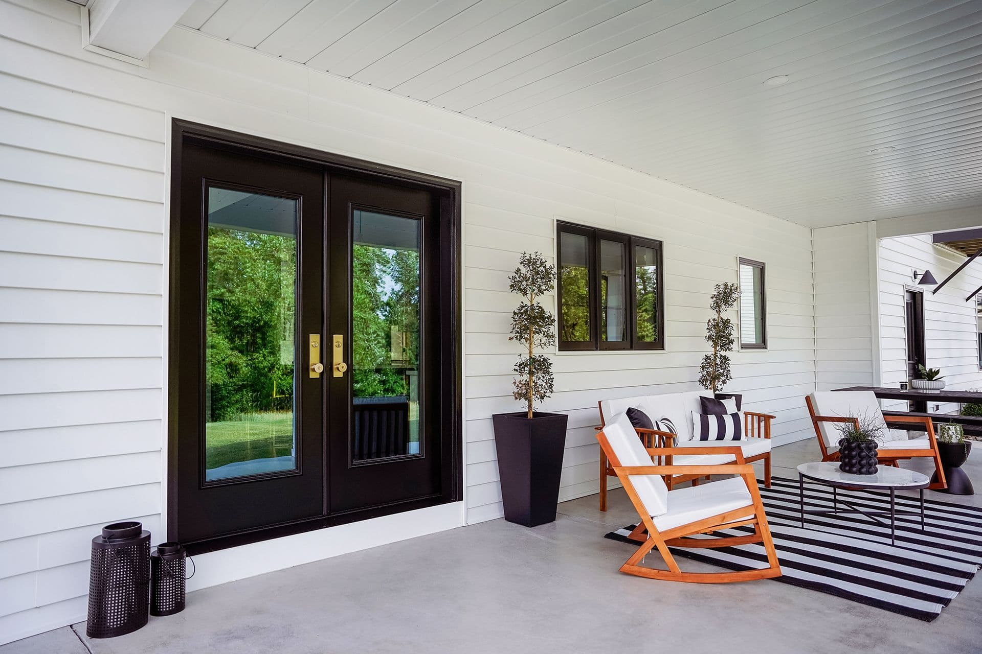 A modern porch with white siding features black double doors, two potted topiaries, wooden chairs with white cushions, a striped rug, and a small table.