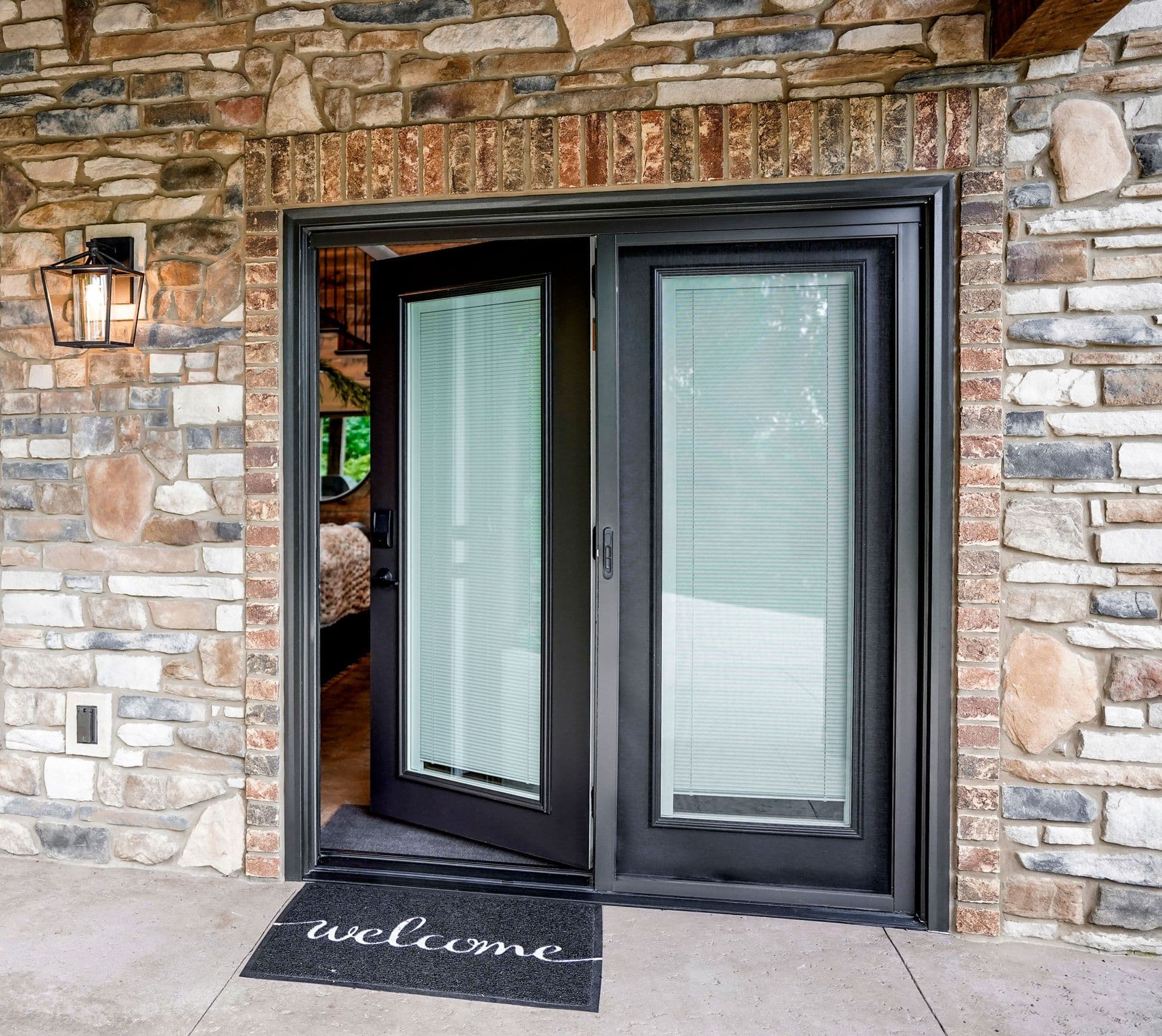 Stone-front entrance with a black-framed glass door, partially open, revealing a cozy interior. A welcoming doormat rests on the concrete step.