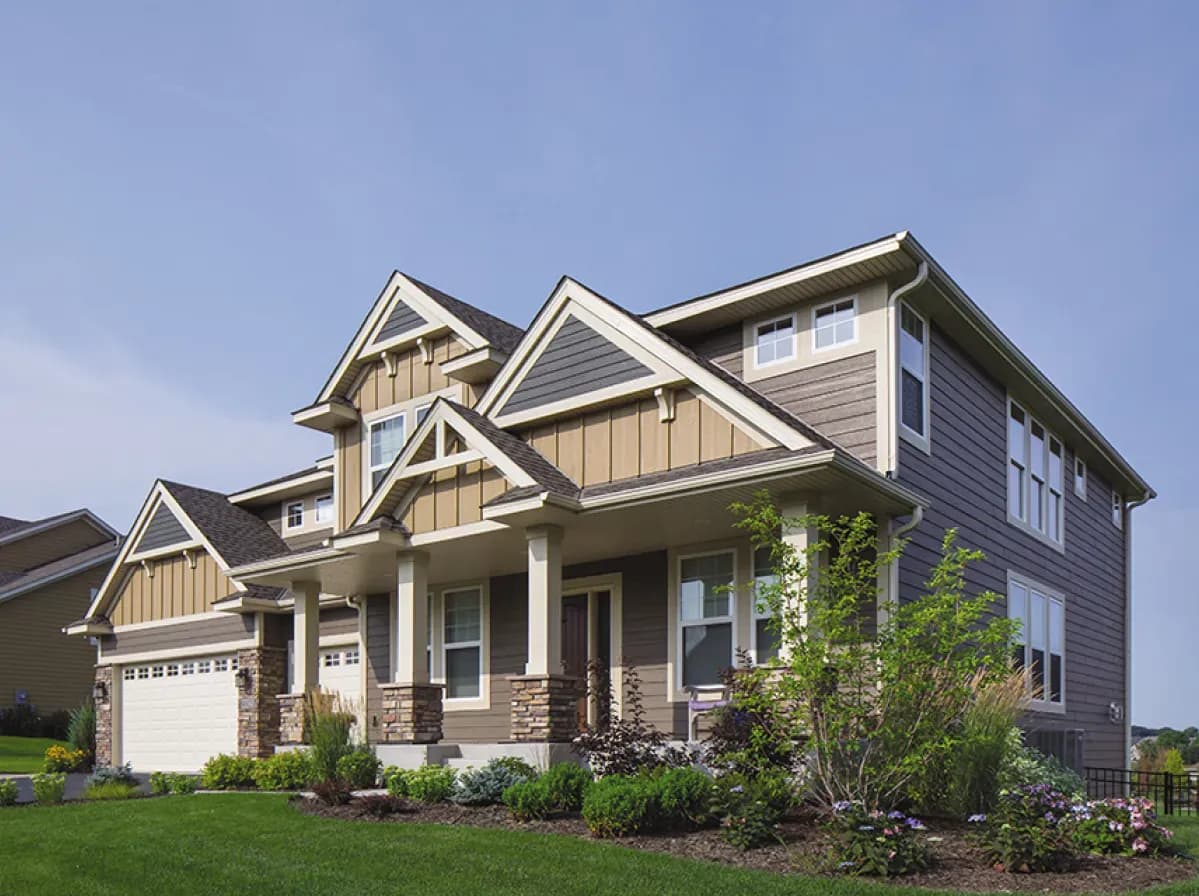 Modern two-story house with beige and gray siding, white trim, and stone accents. Well-maintained garden and lush lawn under a clear blue sky.