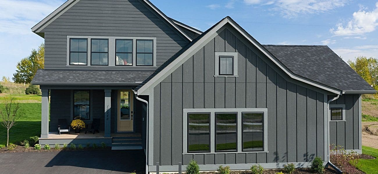 A modern, two-story gray house with large windows is set against a clear blue sky. The home features a manicured lawn and a covered front porch.