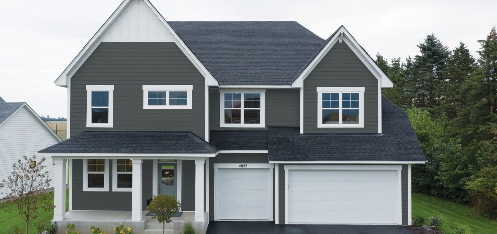 A two-story, modern gray house with white trim, featuring a covered porch and a two-car garage. The setting is suburban with lush green trees.