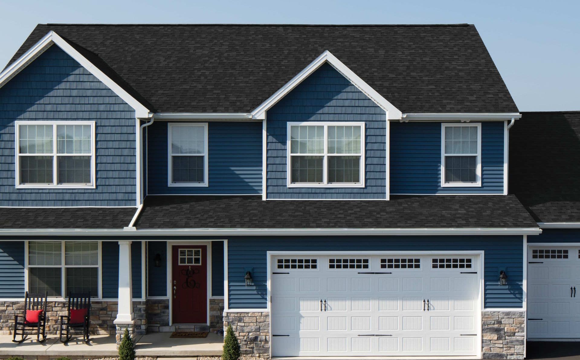 Front exterior of a blue two-story house with dark roof, white trim, red entry door, two-car garage, and stone foundation