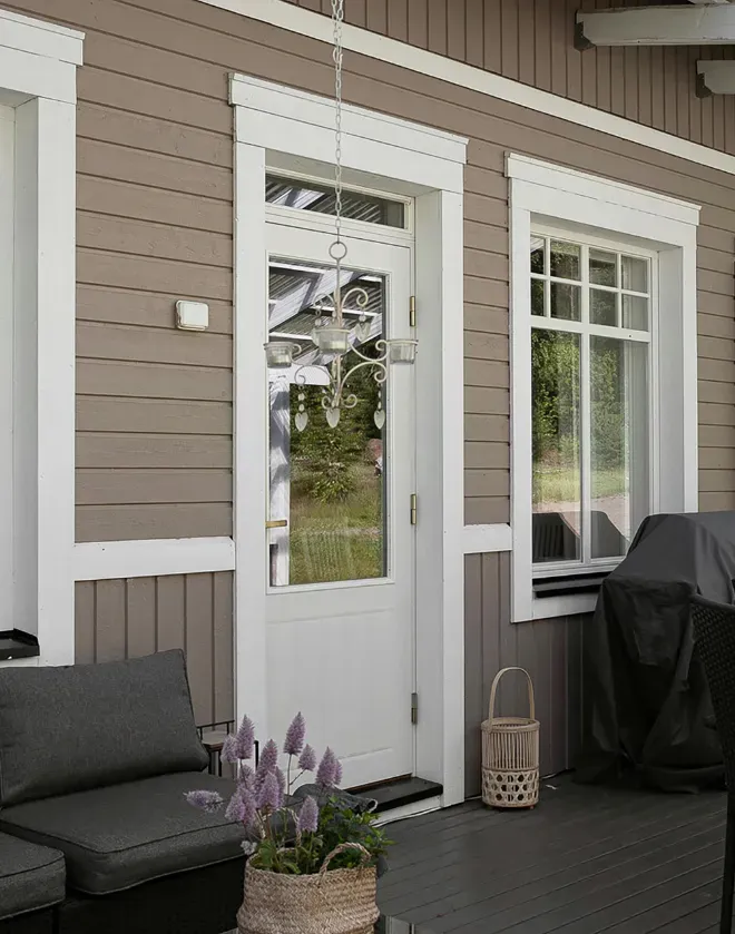 Cozy porch with taupe siding, white trim, and a glass door. A gray sofa, lavender flowers in a basket, and a lantern create a welcoming feel.