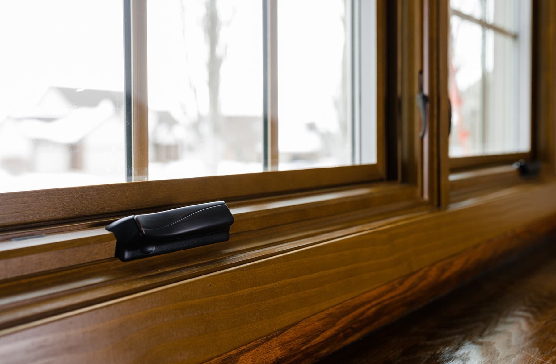 Close-up of a wooden window frame with a black latch, looking out to a snowy neighborhood. The tone is warm and cozy despite the cold outside.