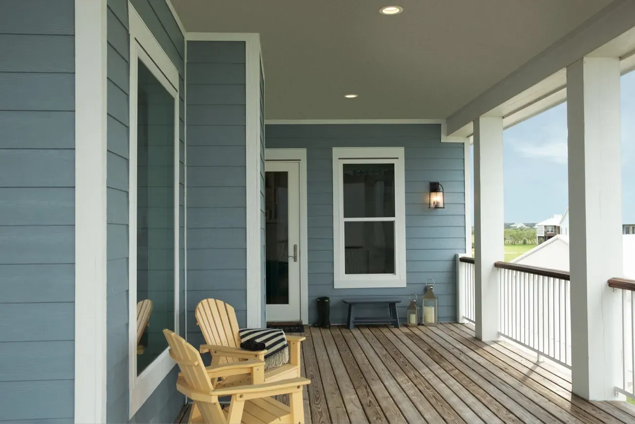 Covered porch with gray-blue siding, wooden floorboards, and a white-framed door and window. A wooden chair nearby suggests a cozy, calm atmosphere.