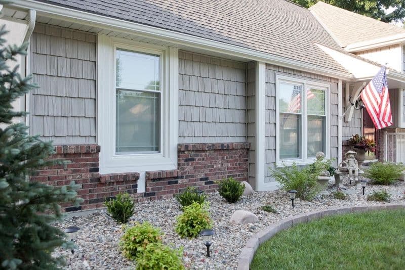 Single-story house with beige siding and red brick accents. Windows are framed in white. An American flag is displayed near the porch, and the garden features small shrubs and decorative rocks.