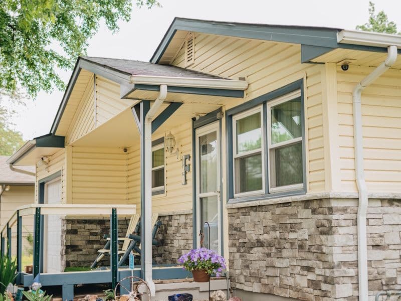 A cozy, single-story house with yellow siding, stone facade, and gray trim. A small porch features a potted plant with purple flowers, evoking a welcoming feel.