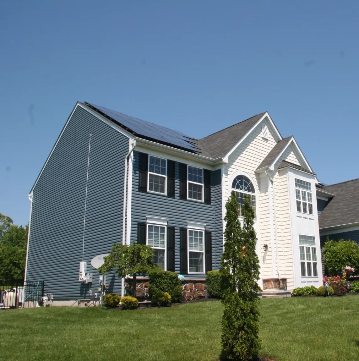Two-story house with blue and white siding, solar panels on the roof, and large windows. The front lawn is green with trimmed bushes under a clear blue sky.