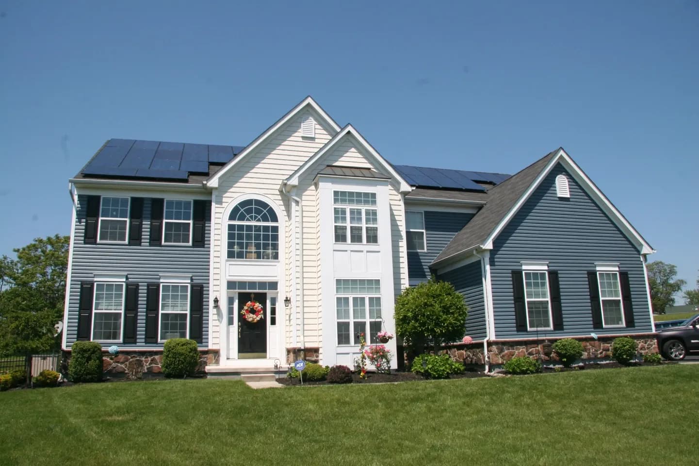 Two-story blue and white house with solar panels and a wreath on the front door. Surrounded by a lush green lawn and sunny blue sky.