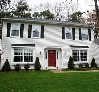 A two-story white colonial house with a red door, black shutters, large windows, and neatly trimmed shrubs on a lush green lawn. Overcast sky in the background.