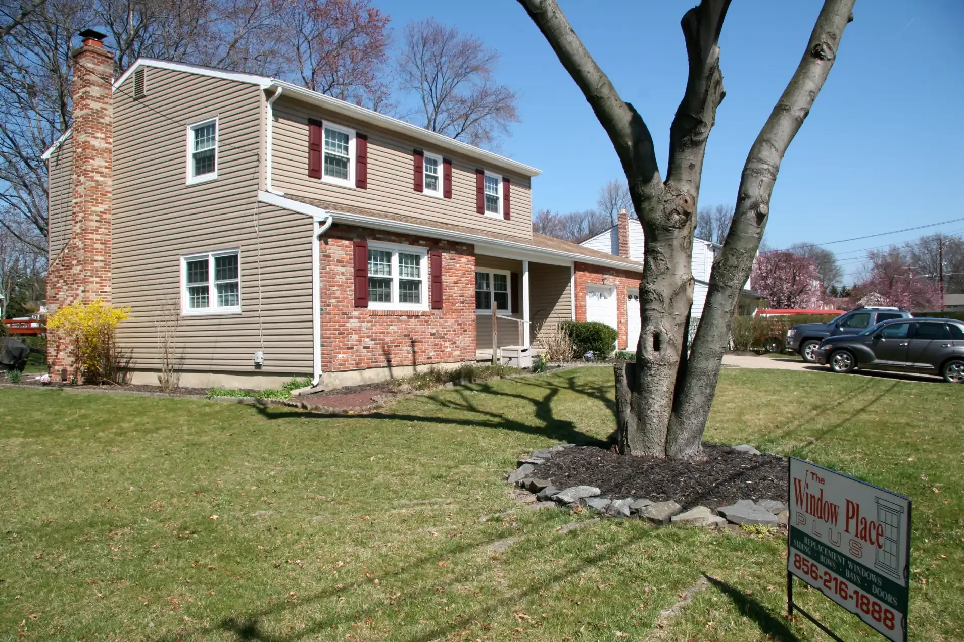 Two-story suburban house with beige siding and red brick facade, set in a grassy yard with a bare tree. A sign for a window company is in the foreground.
