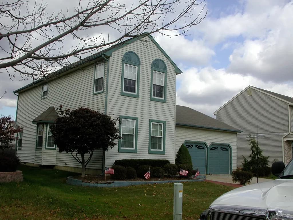 Two-story house with teal-trimmed windows and garage, surrounded by small trees and bushes. American flags line the yard. Overcast sky.