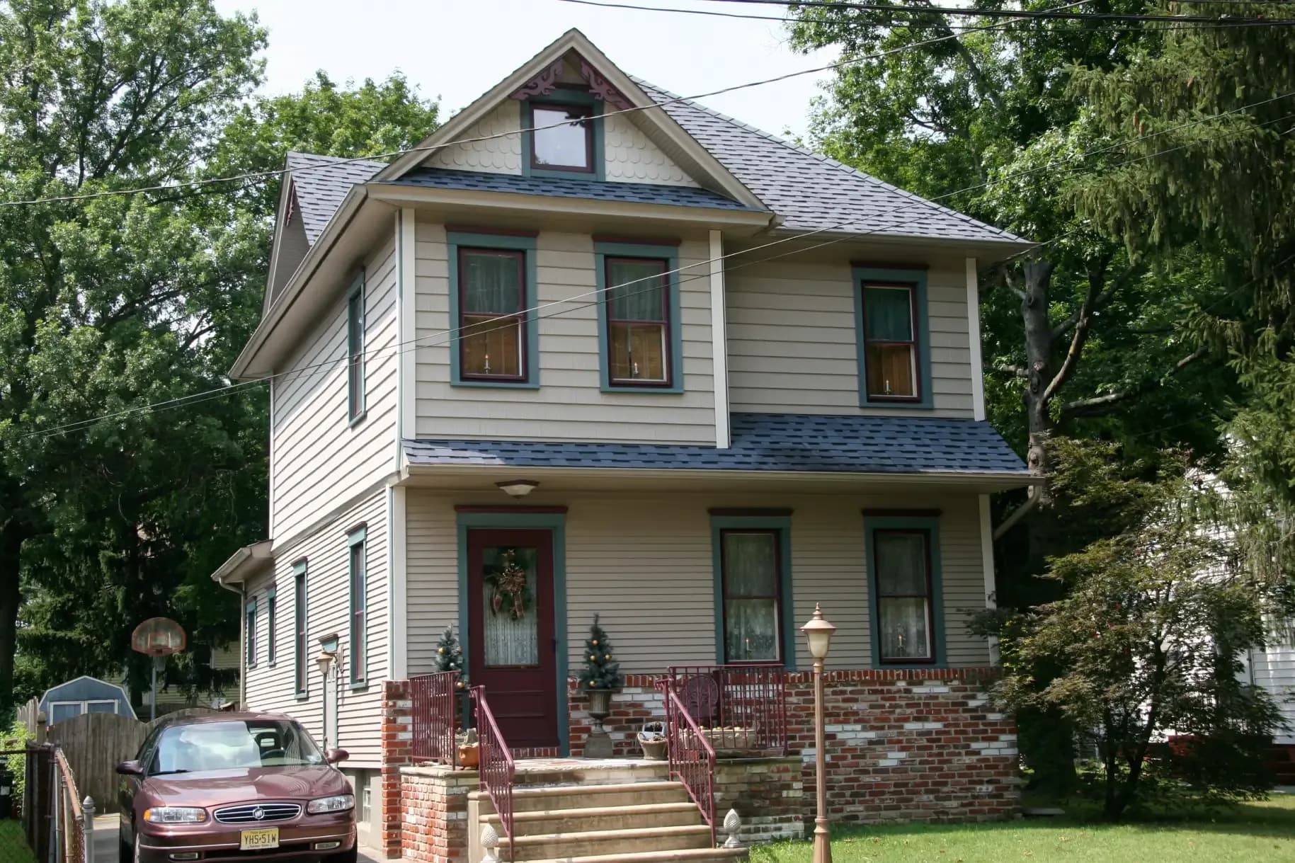 A two-story suburban house with beige siding and a dark roof. It features a red front door, a small porch with steps, and a car parked in the driveway.