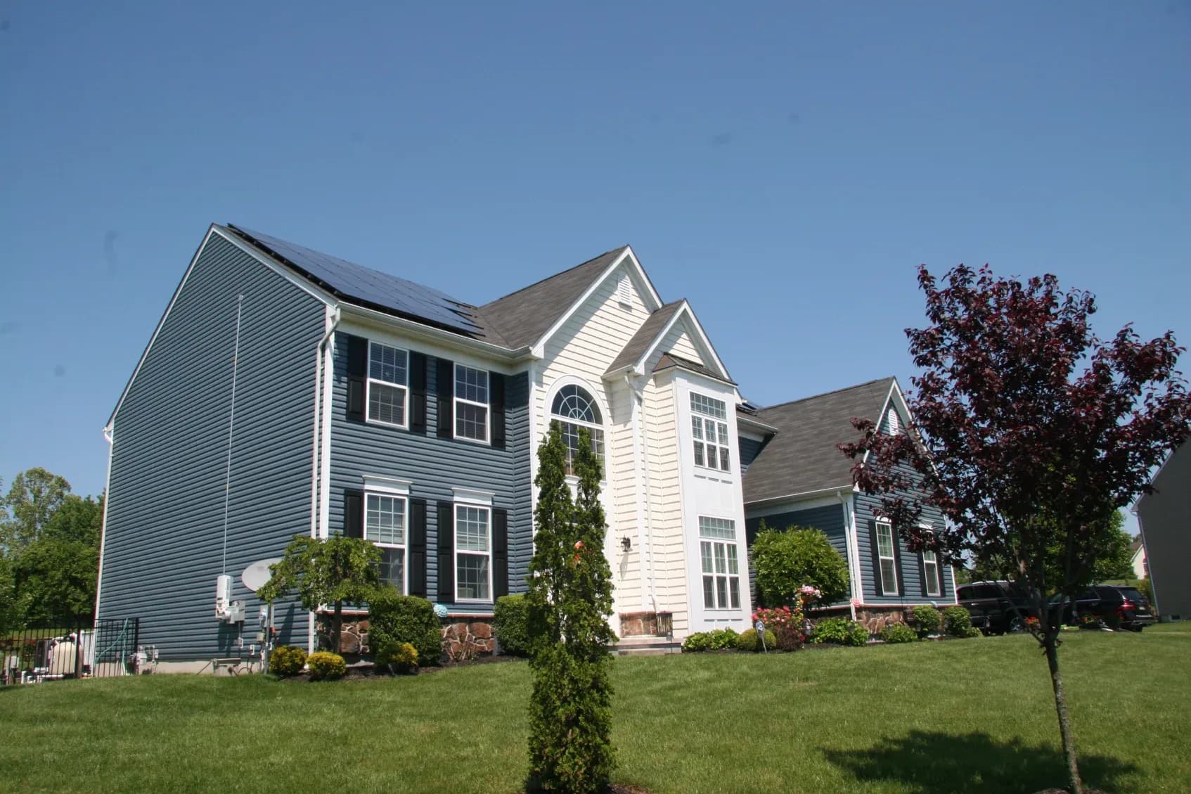 Large, two-story blue and white suburban house with solar panels on roof, surrounded by a well-maintained lawn and trees set against a clear sky.