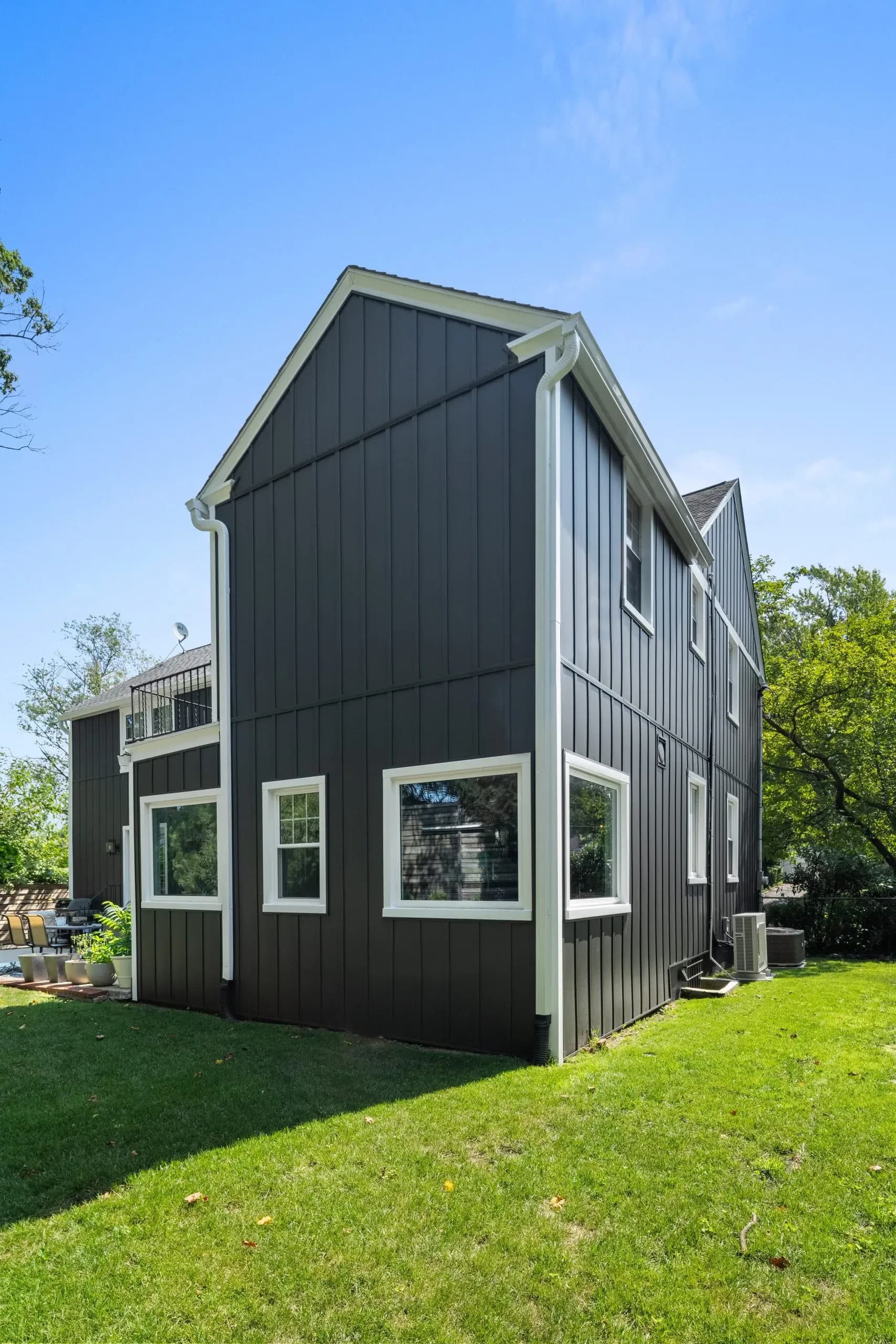 Two-story house with dark siding and white trim, surrounded by lush green grass and trees against a clear blue sky. Modern and serene atmosphere.