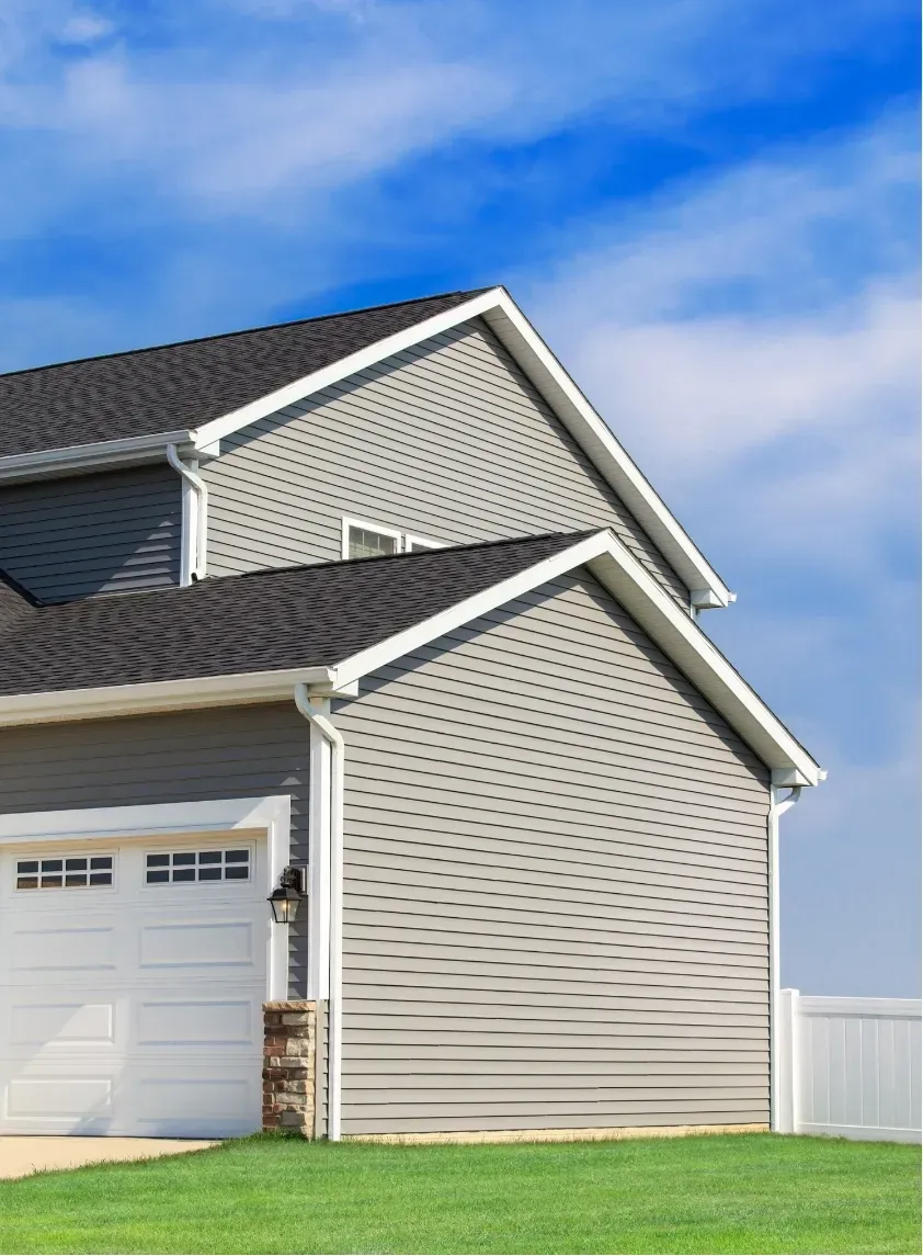 A modern gray house with white trim, a black shingled roof, and a white garage door stands against a blue sky. The yard is a vibrant green.