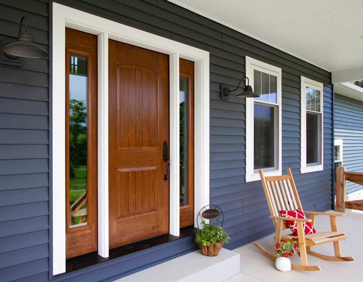A cozy porch with a wooden door framed by navy siding and white trim. A wooden rocking chair with a red pillow sits beside a welcome sign and potted plant, radiating warmth.