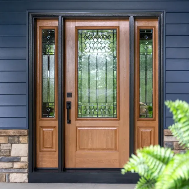 A wooden front door with intricate glass panels, flanked by sidelights, set against dark blue siding and stone accents. A leafy plant is in the foreground.