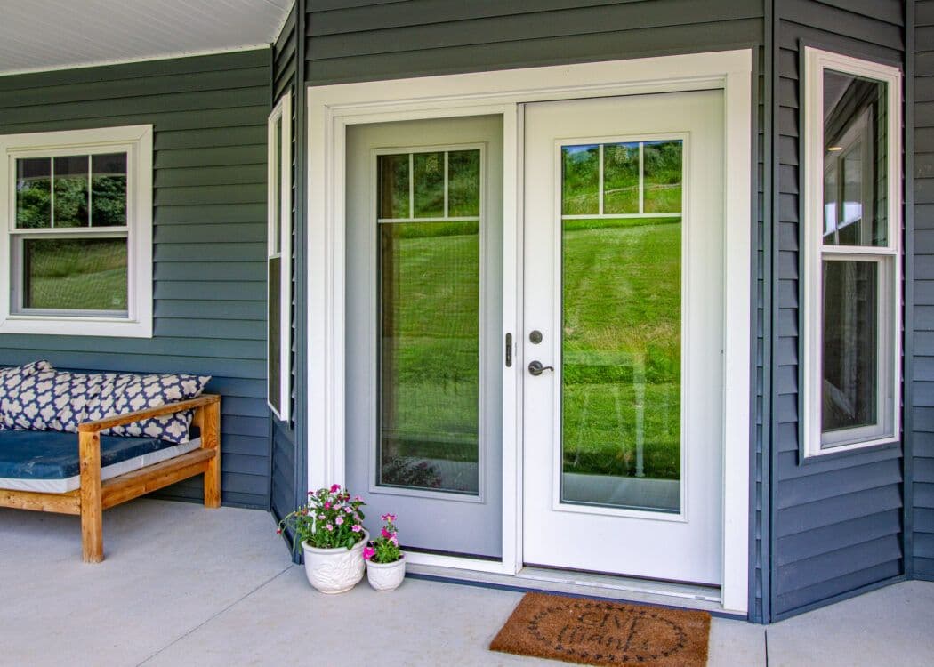 White patio door with glass panels on a dark blue house. A wooden bench with patterned cushions sits to the left. Two potted flowers and a doormat are in front.