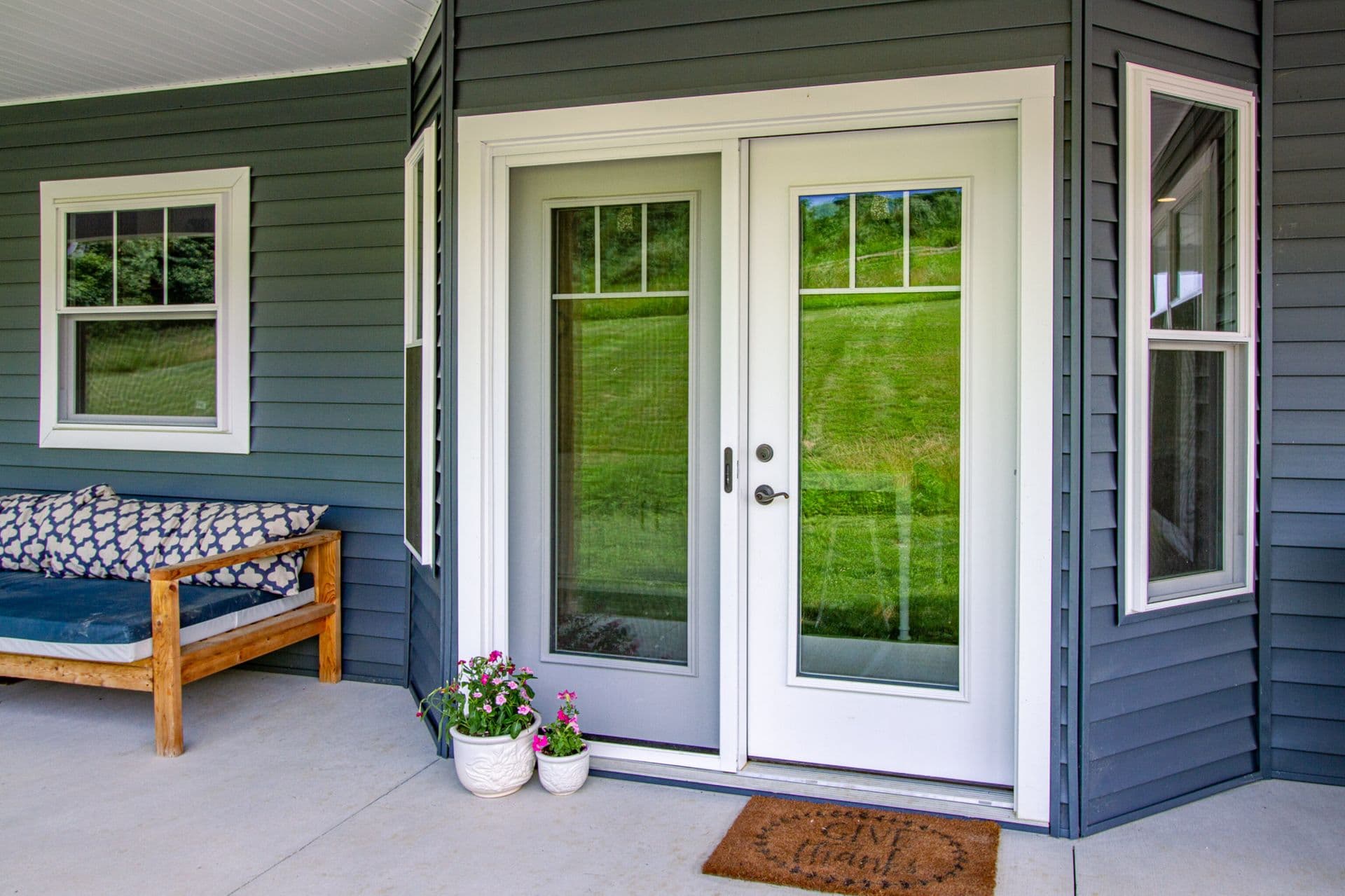 Cozy porch with a blue bench, floral cushions, and double glass doors. Potted flowers add a welcoming touch. The scene is calm and inviting.