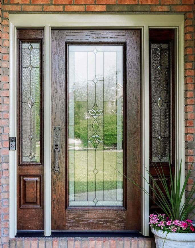 Wooden front door with glass panels etched in a geometric pattern. Framed by brick walls, a potted plant with flowers and green foliage sits to the right.