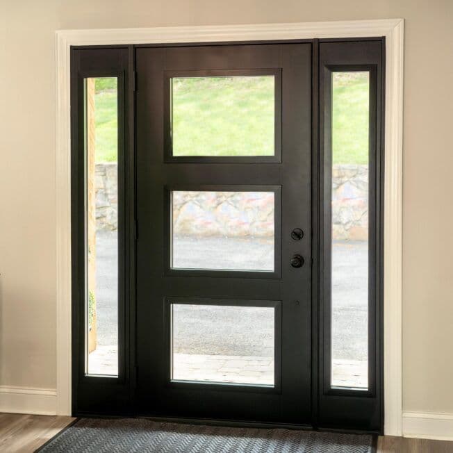 Modern black front door with three vertical glass panes, flanked by slim side windows. Sunlight filters through, casting soft shadows inside.