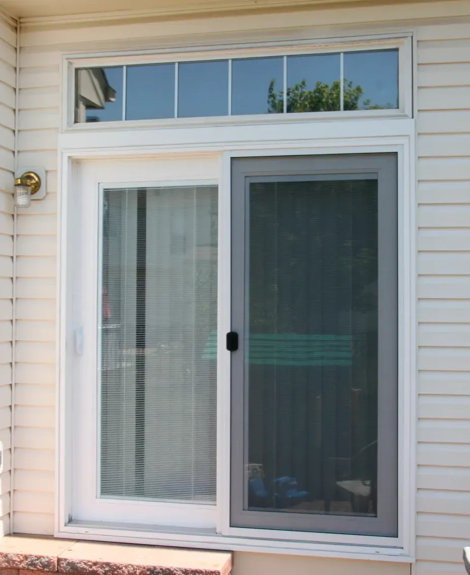 Sliding glass patio door with vertical blinds and a mesh screen on the right. Above, there is a row of small horizontal windows. The siding is light-colored.