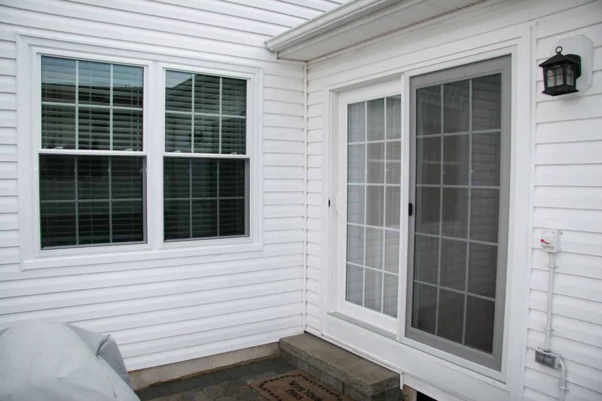 A white house exterior with a window and sliding glass door. The window has grilles, and the door opens onto concrete steps with a welcome mat. A black outdoor lantern is mounted on the wall, creating a clean and inviting entrance.