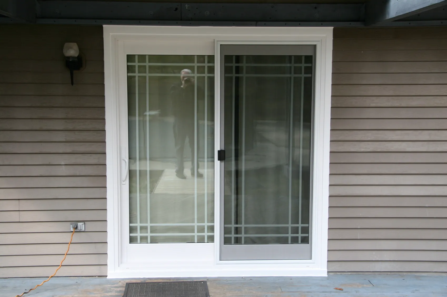 Sliding glass door with white frame, set against beige siding. The door features a grid pattern and reflects the silhouette of a person taking the photo.