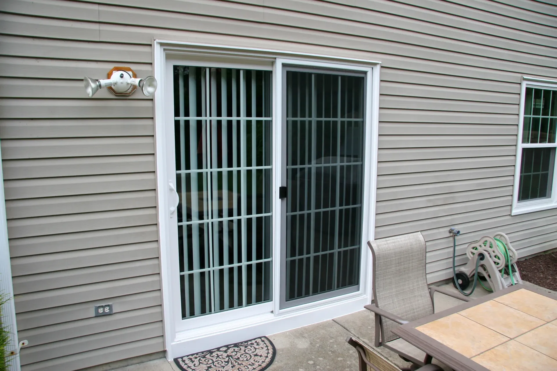A sliding glass door with white blinds is set in a beige vinyl-sided wall. Nearby is a patio table with chairs and a garden hose coiled on a reel.