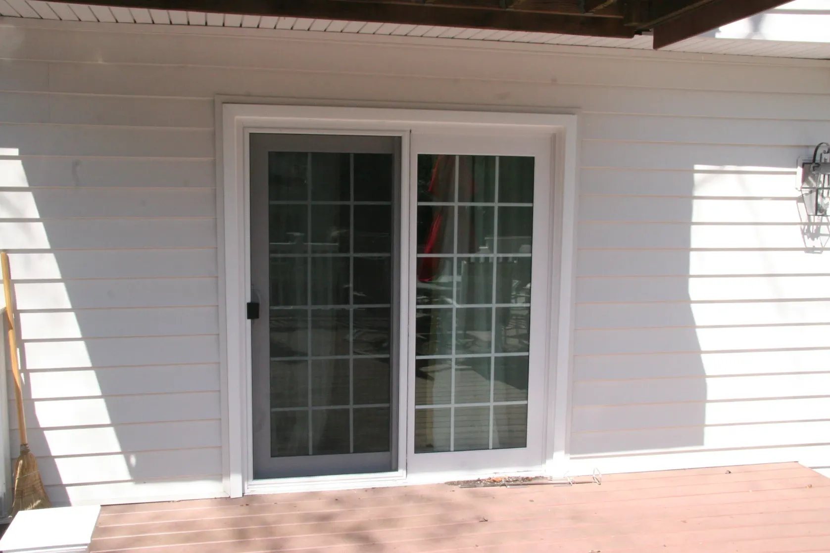 Sliding glass door with white frame on a house with white siding. Sunlight creates shadows on the wooden deck, and a wall light is visible on the right.