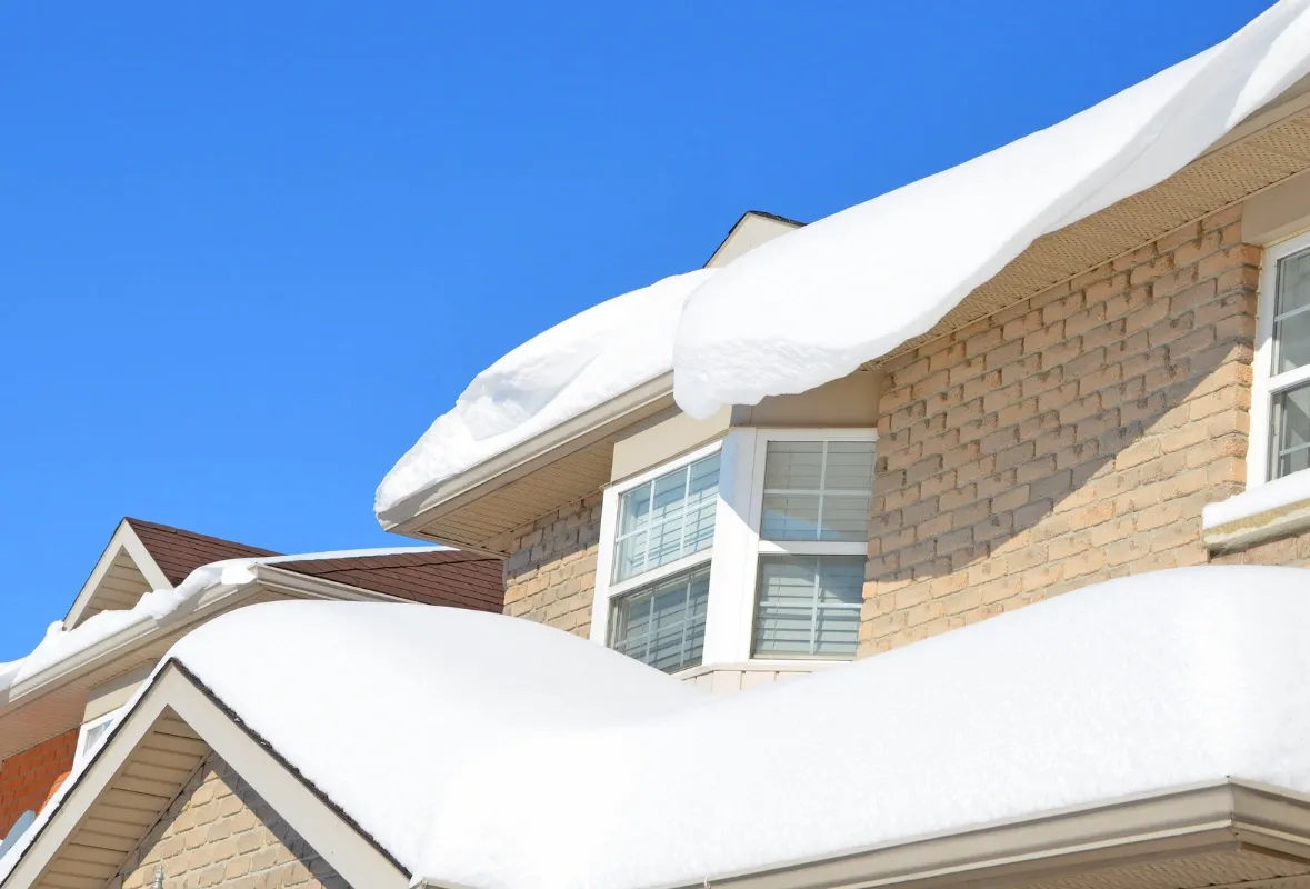 Snow blankets the roof of a brick house under a bright blue sky. The heavy snow creates a peaceful winter scene, highlighting the house's windows and eaves.